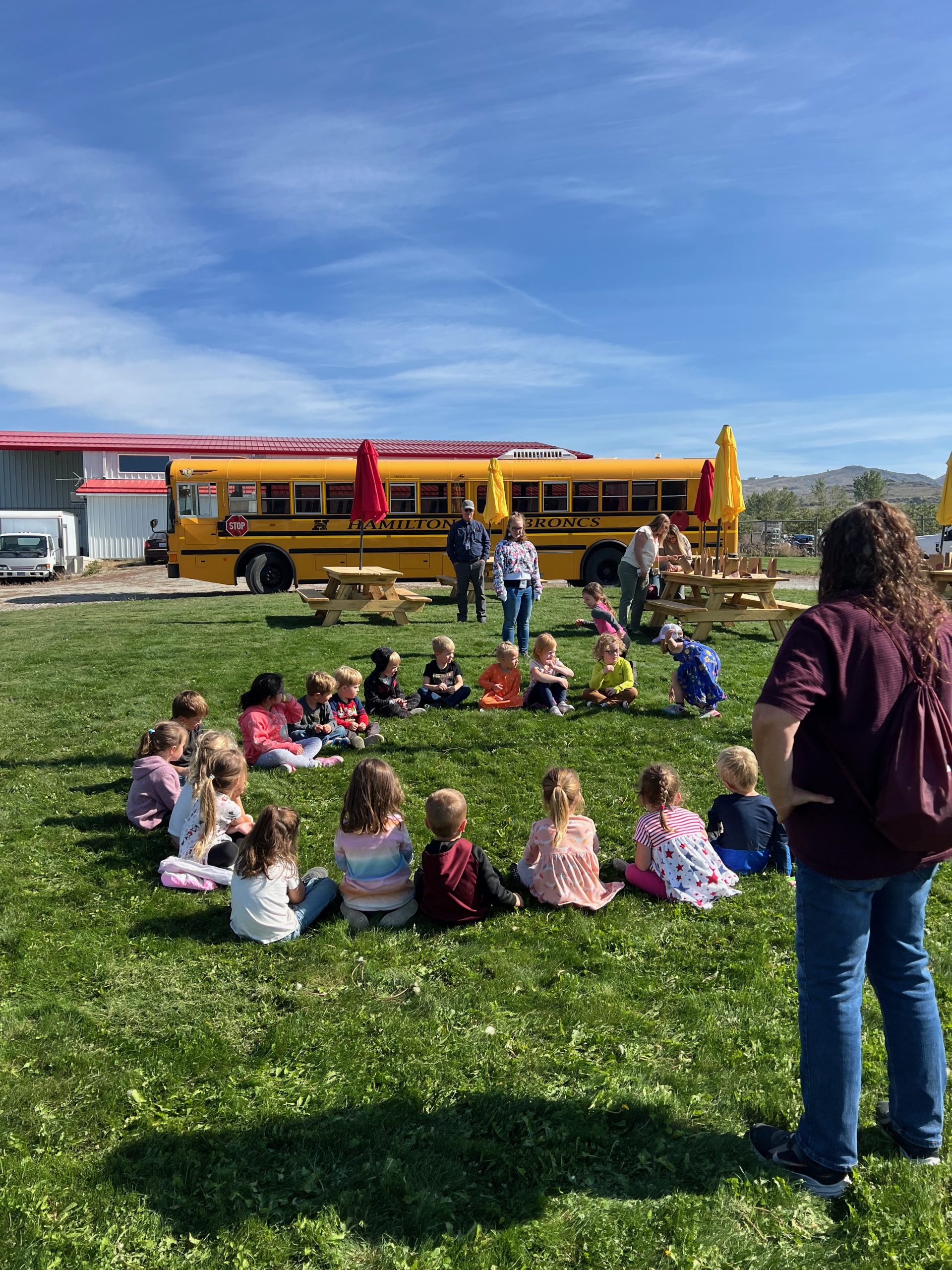 Children sitting in a circle on the grass, with a school bus and the Swanson's Mountain View Orchard packing shed in he back