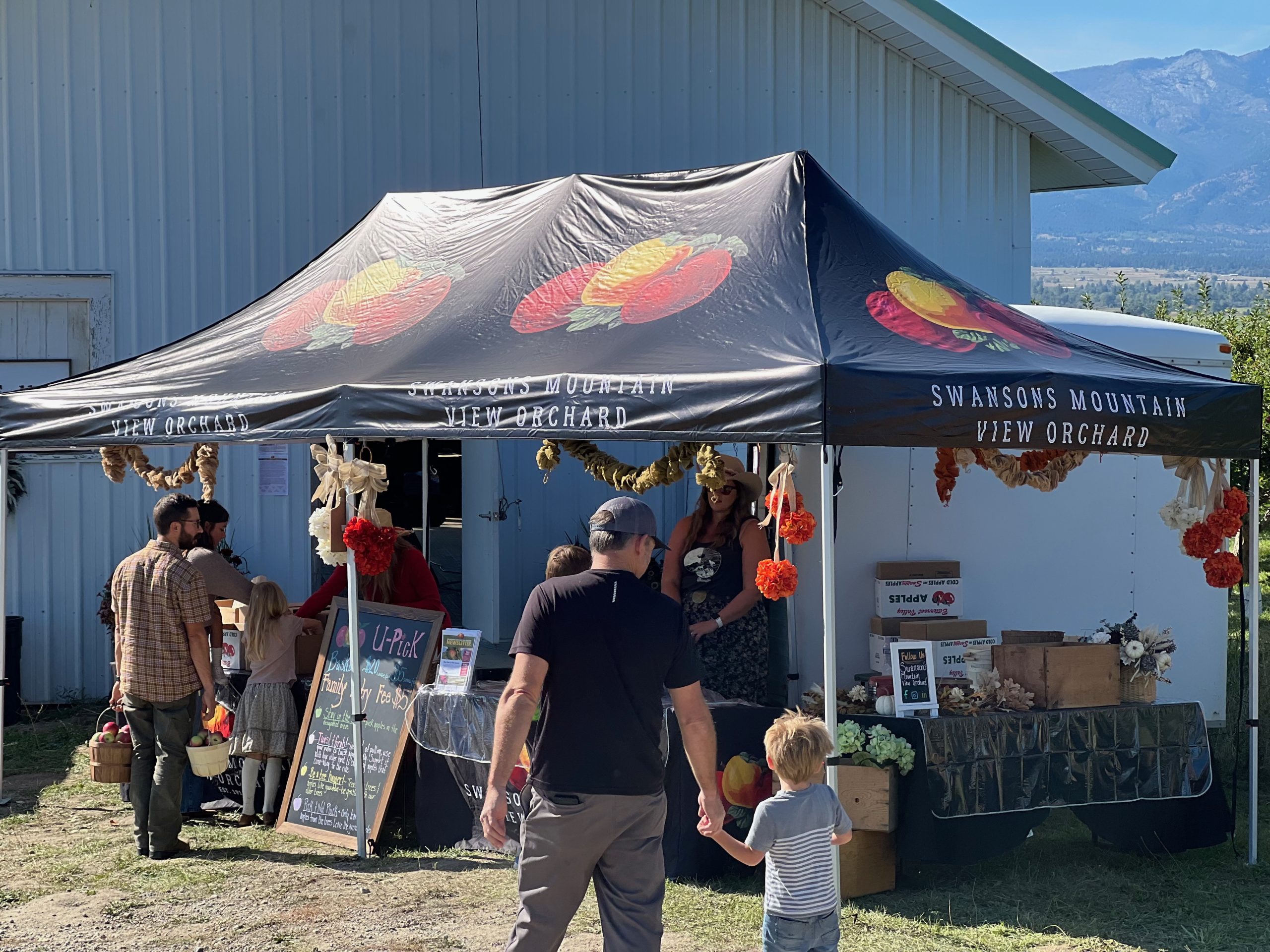 Swanson’s Mountain View Orchard U-Pick tent with customers, staff, fall decorations, and chalkboard sign in Montana