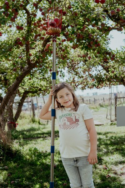 Girl holding apple picker, smiling in orchard in Swanson's t-shirt